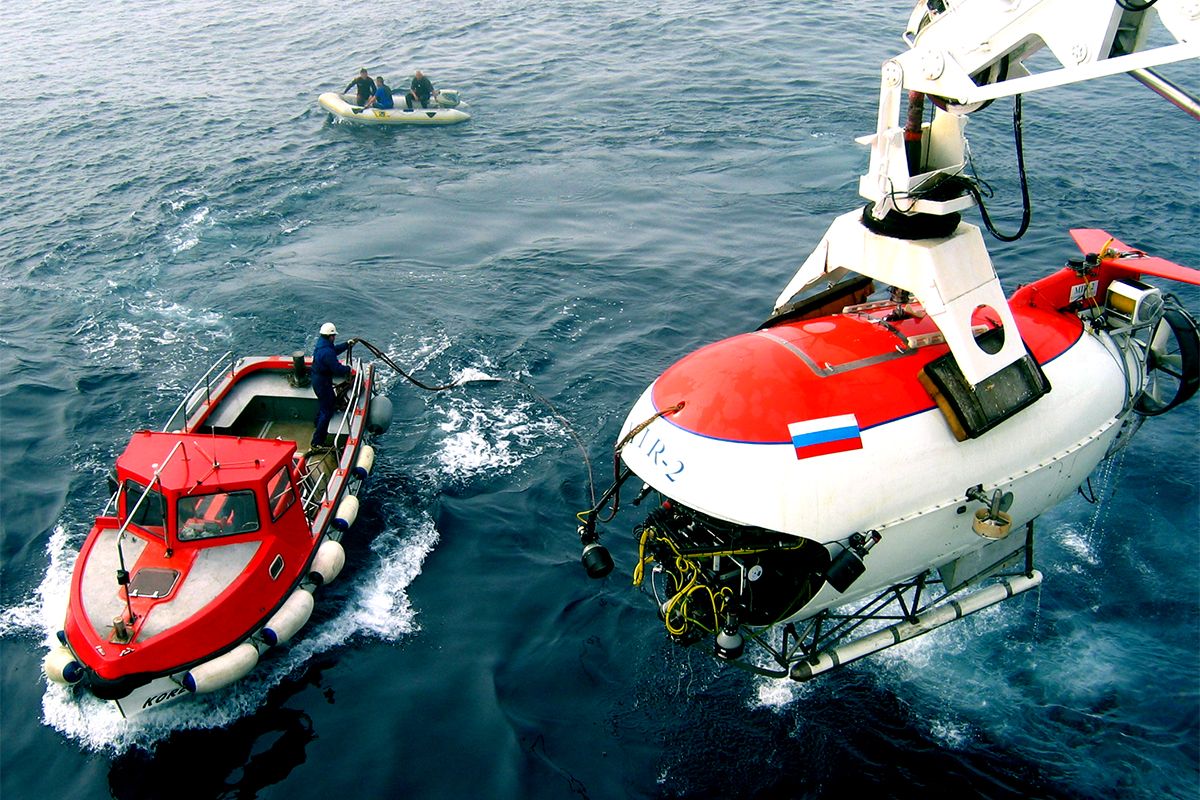 Photo showing a red and white submersible being lowered into the water by a crane. There are also two small boats already in the water.