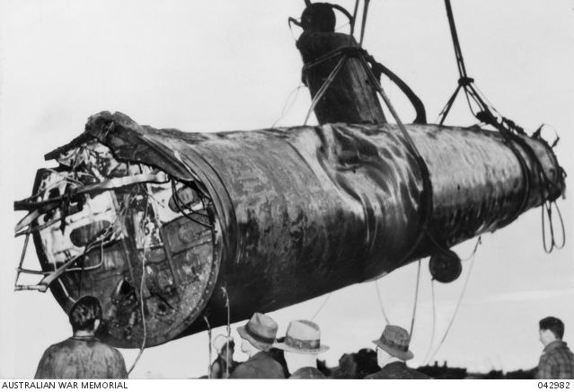 Black and white photo showing a small, damaged submarine being suspended, presumably by a crane.