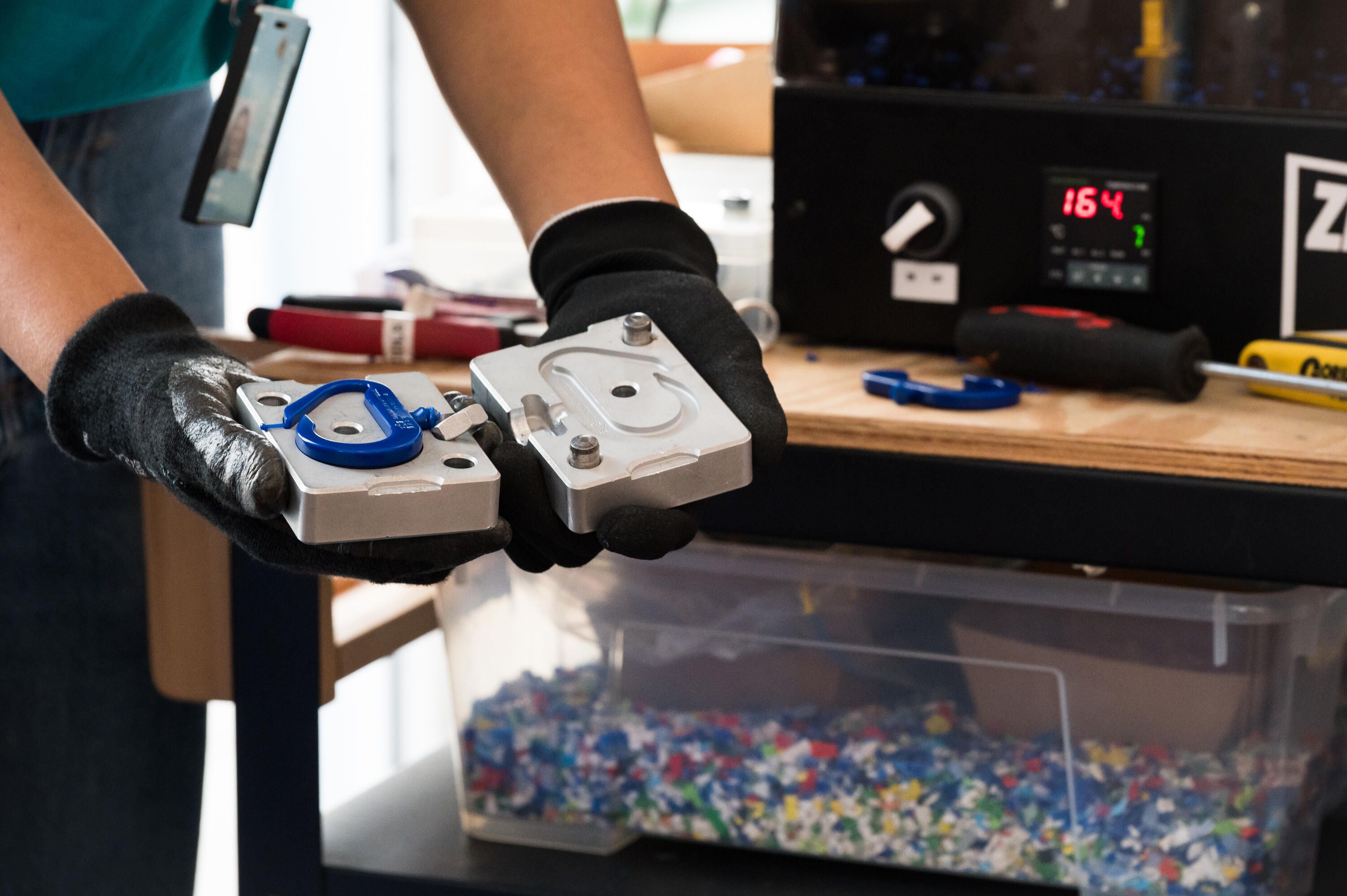 Photo showing a close up of a person with gloves holding a metal mould with a blue plastic carabiner being made in it.