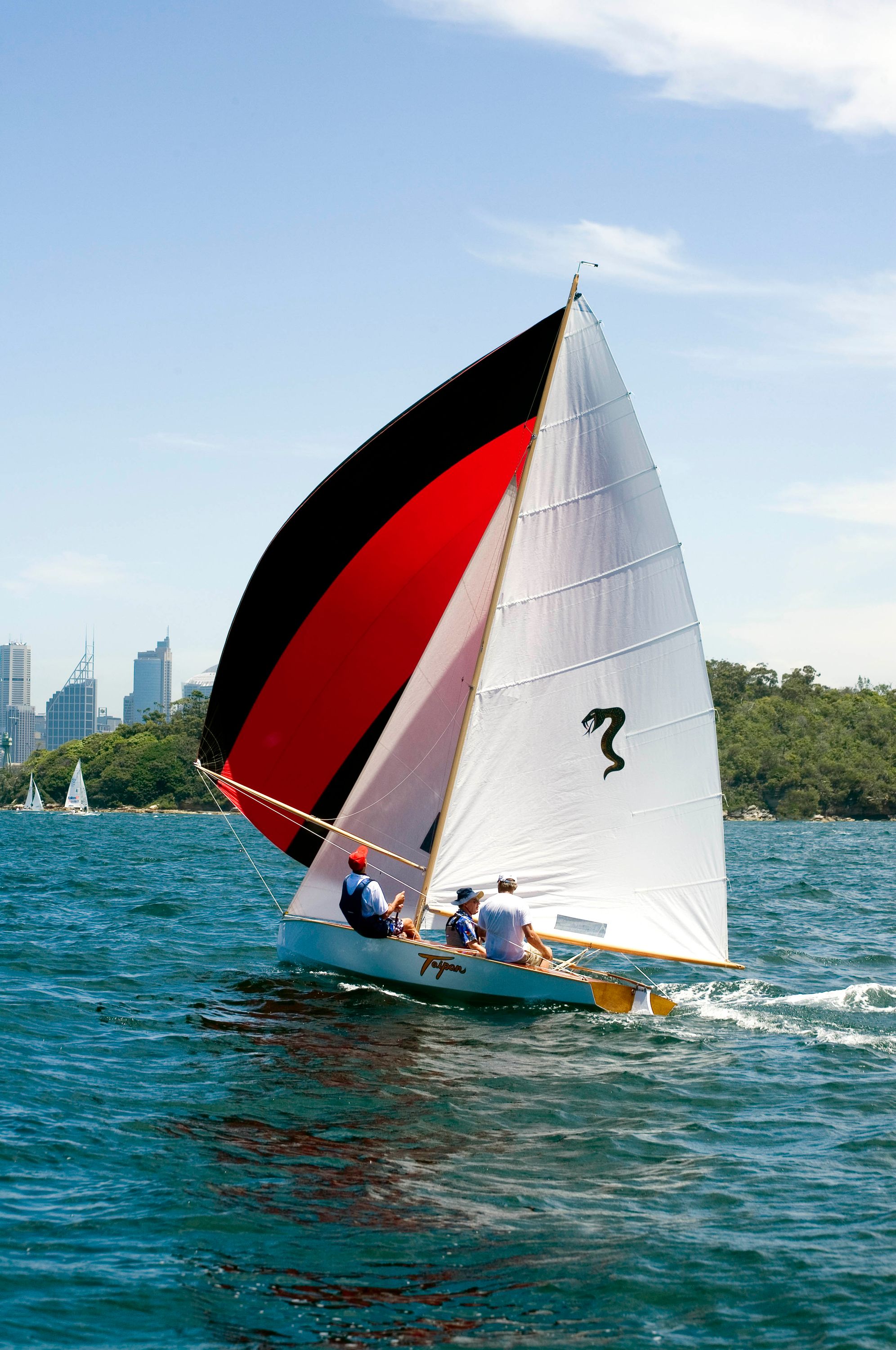 Colour photograph showing a sailing boat with white, red and black sails, with 3 crew sailing it.