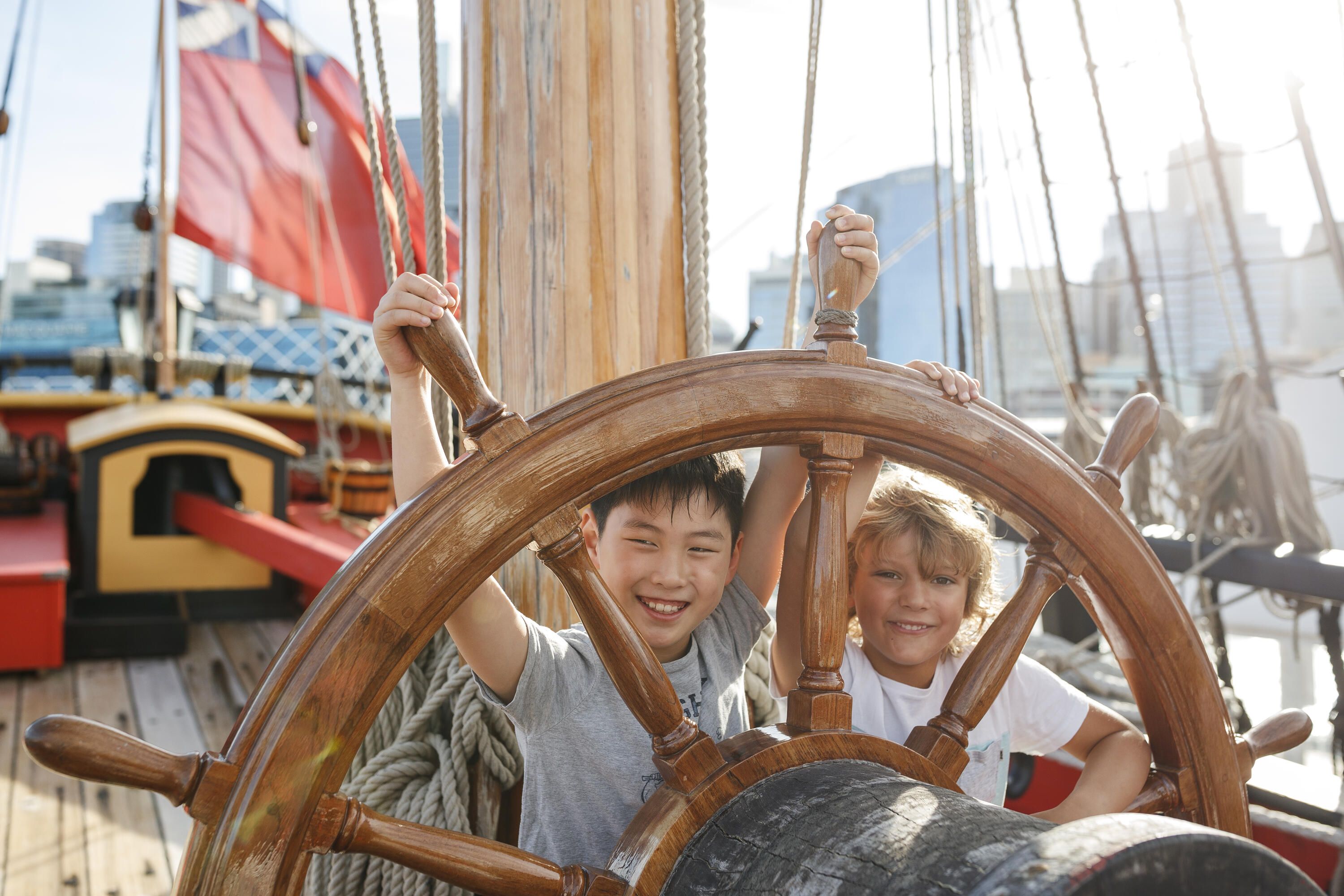 Photo showing Two young kids (boys) stand behind the wheel on a wooden tall ship.
