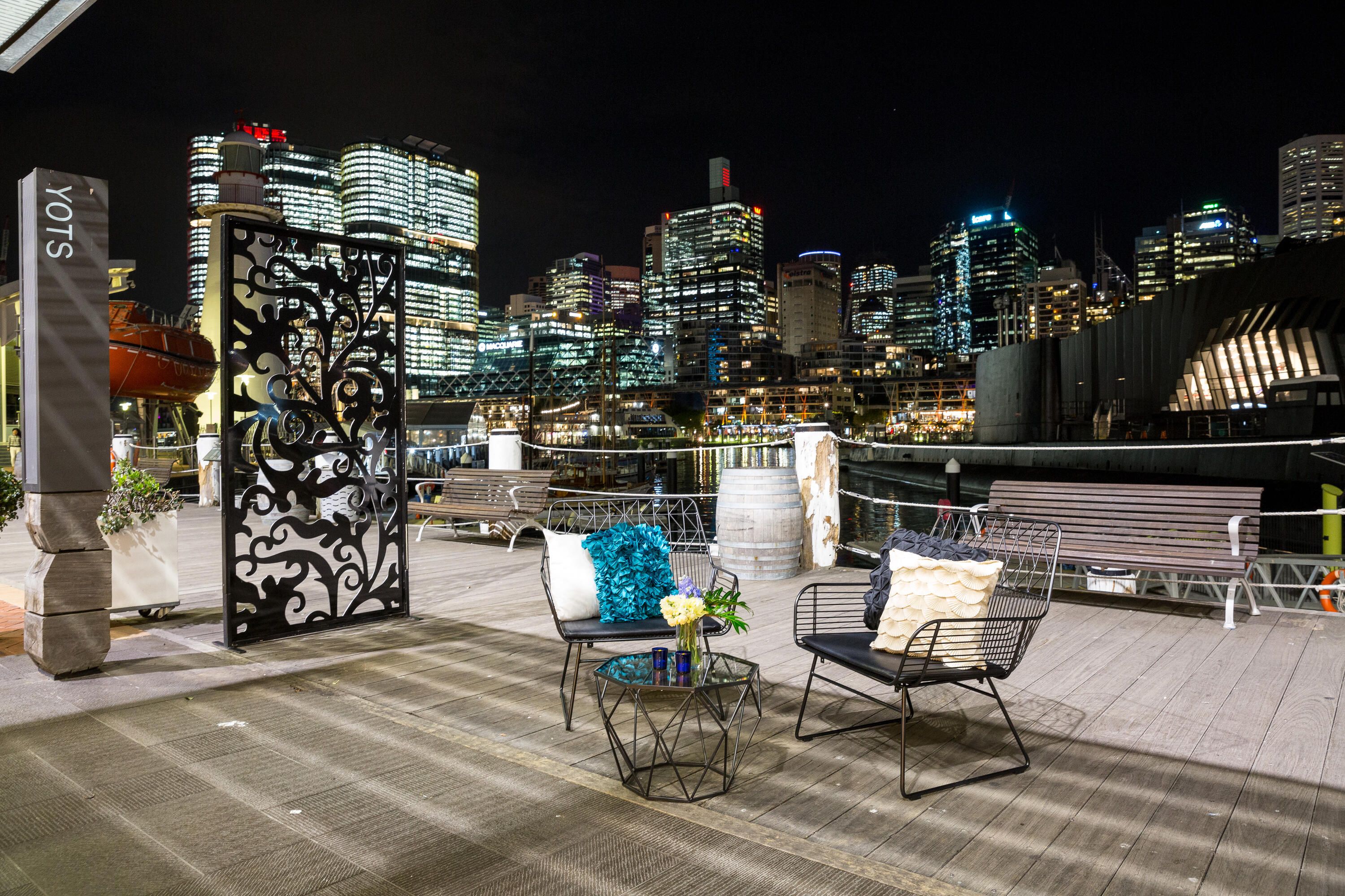 photo taken at night of boardwalk with outdoor furniture and Sydney skyline in the background. 