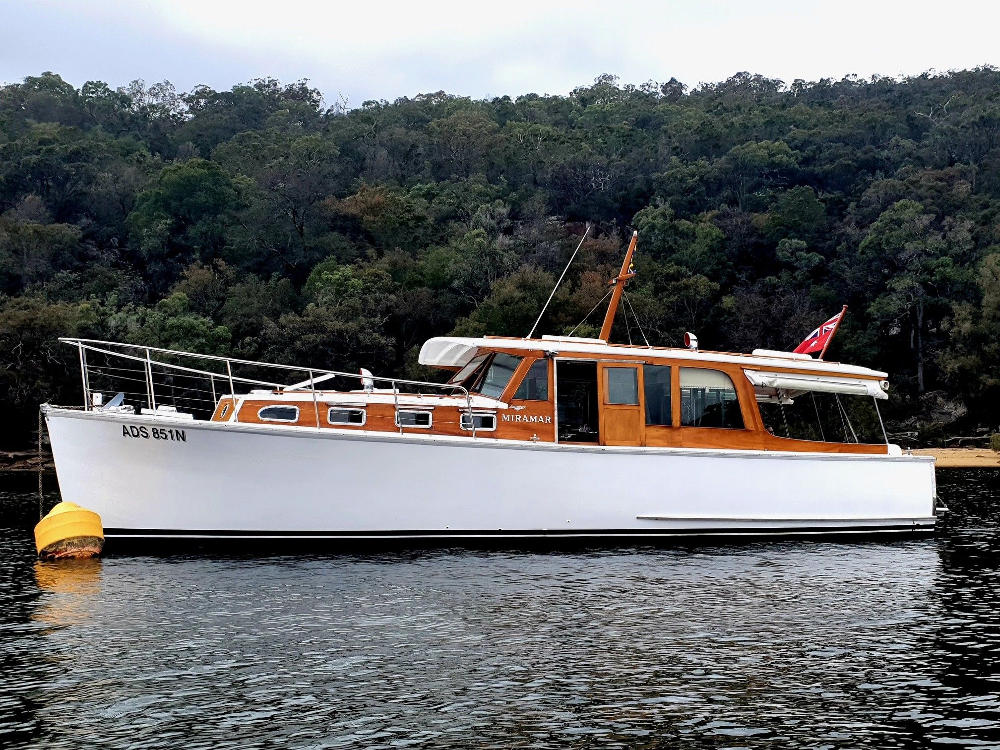 Photo of a white and wood boat floating on the water, with dark green foliage behind it. 