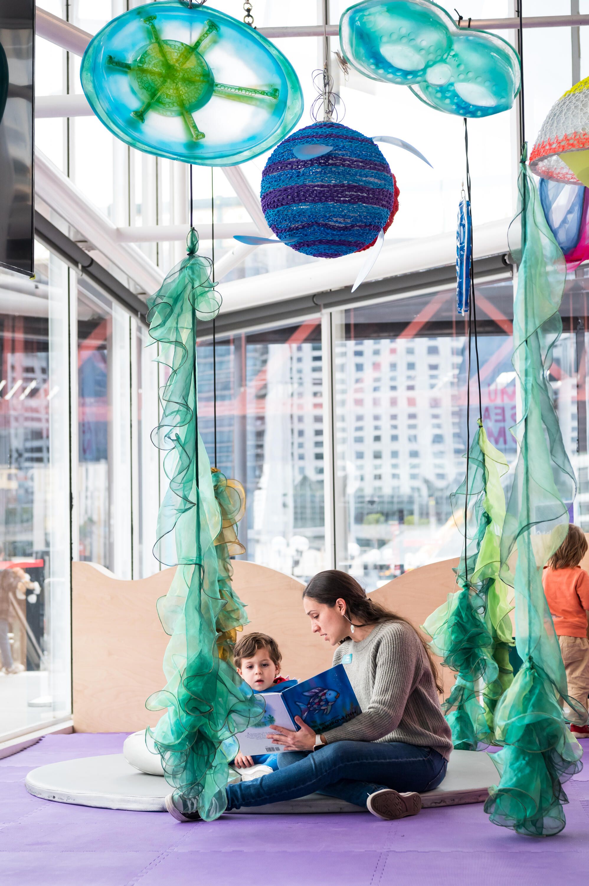 A woman and young boy reading a book in a bright space with hanging, sea themed decorations.