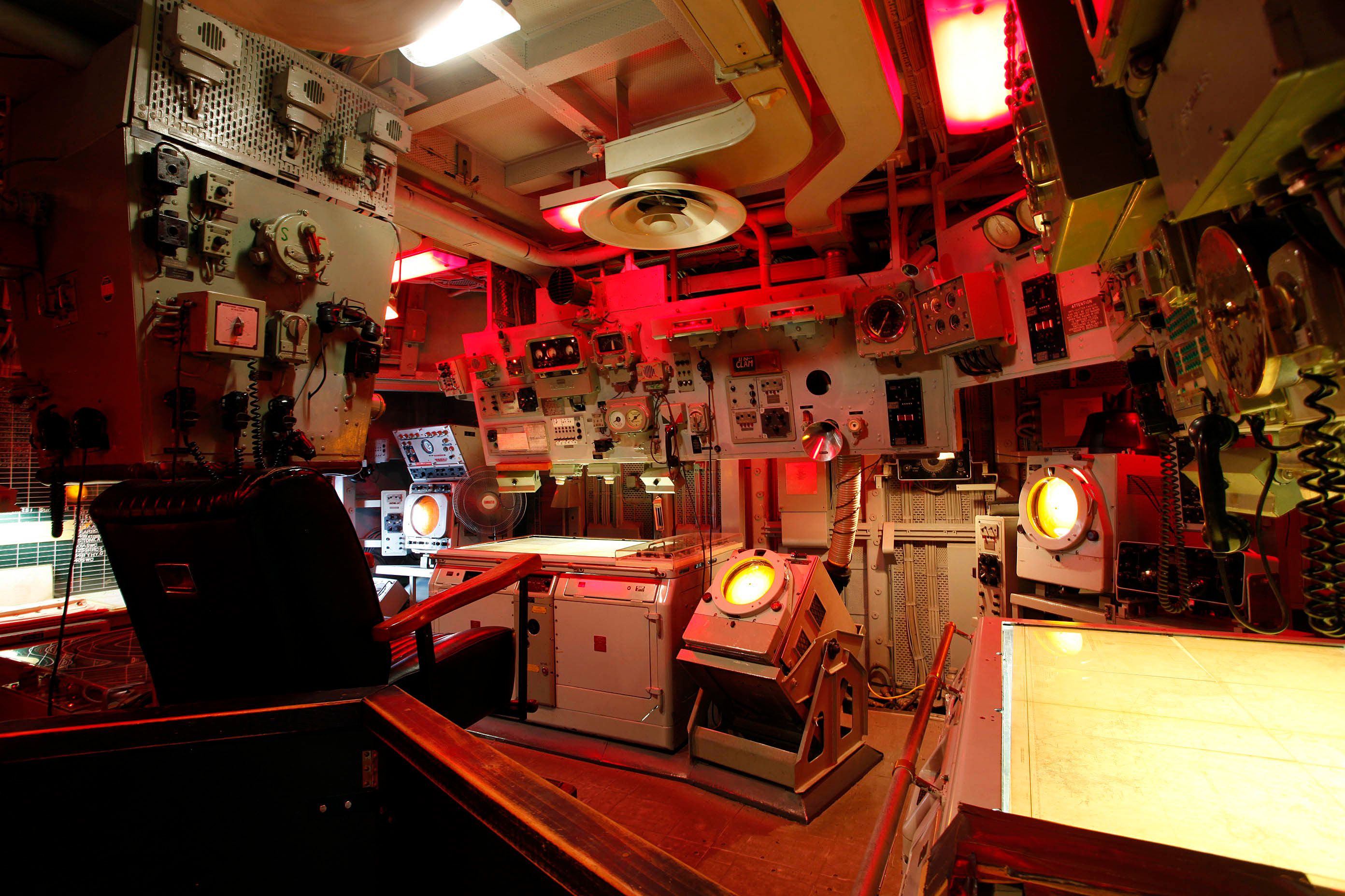 Photo showing a room on a navy ship, with navigation equipment and red lighting. 