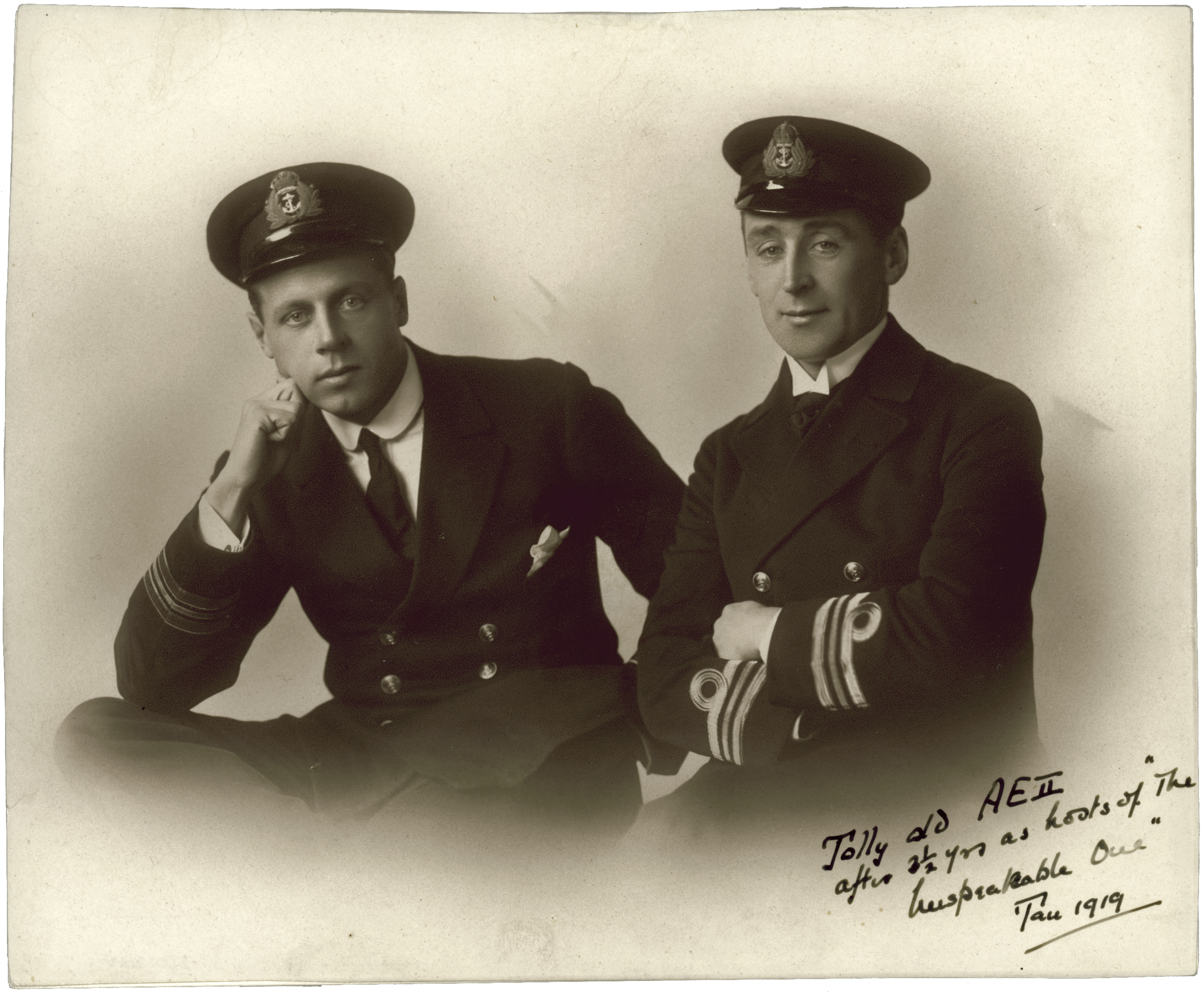 Black and white portrait photograph of two men in uniform including hats featuring an anchor badge. Written on the bottom right corner in cursive writing is "Jolly old AE2 after 3 1/2 yrs as hosts of "The Unspeakable One" Jan 1919". 