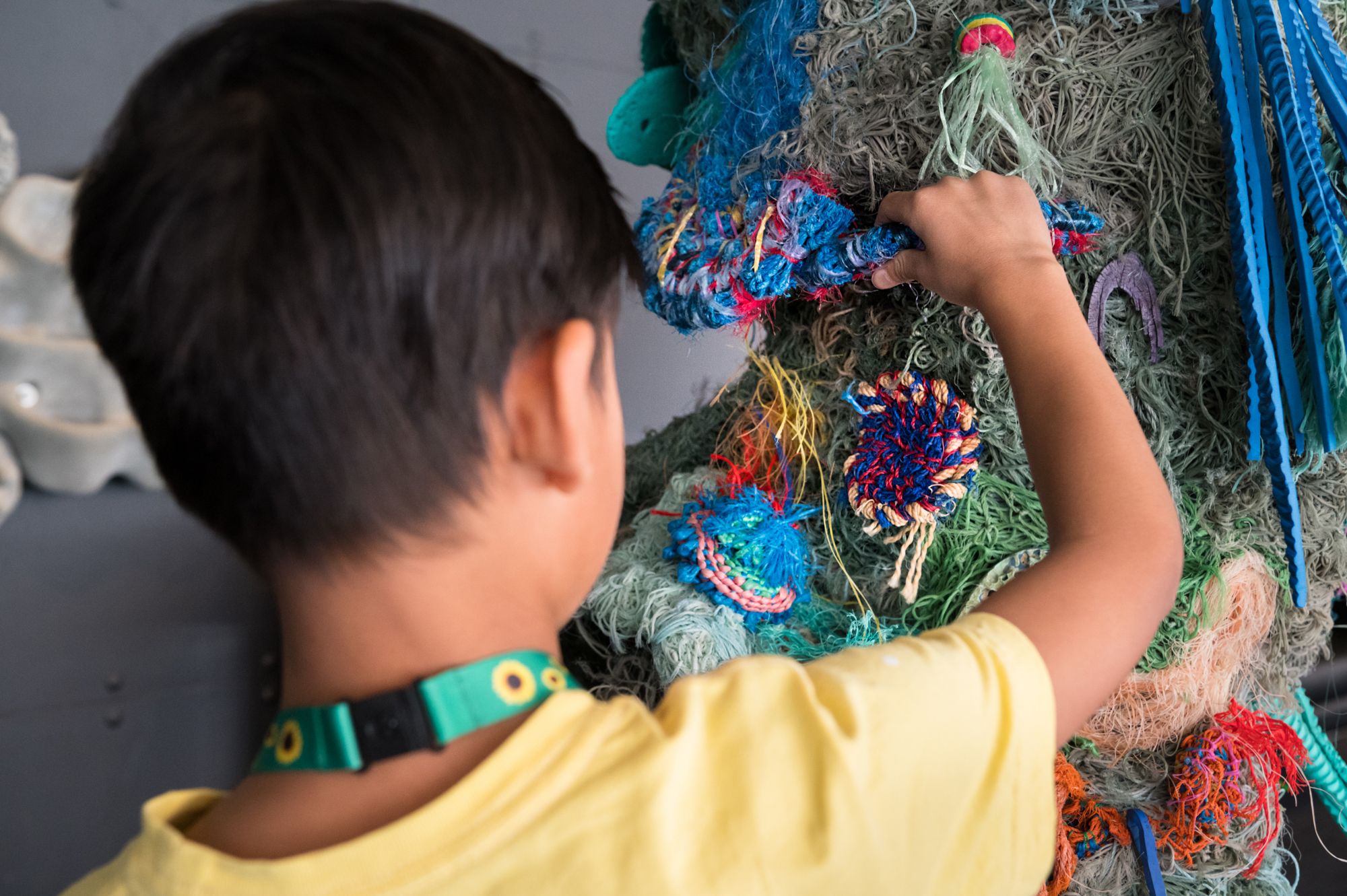 Young boy with short, dark hair, wearing a yellow shirt and sunflower lanyard touching a coral reef sculpture made of nets