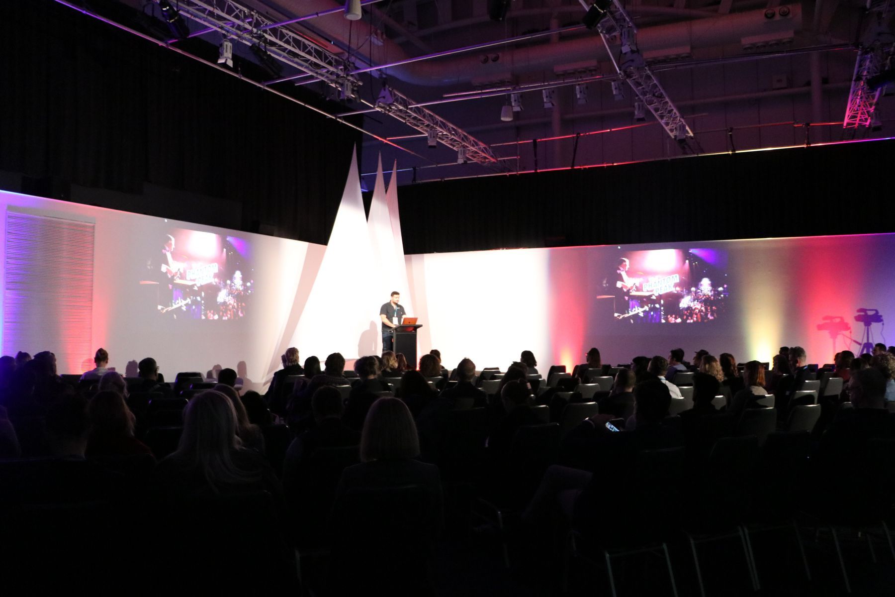 Photo of a museum event space set up like a threatre, with a large numper of eated guests looking towards a presenter on a stage.