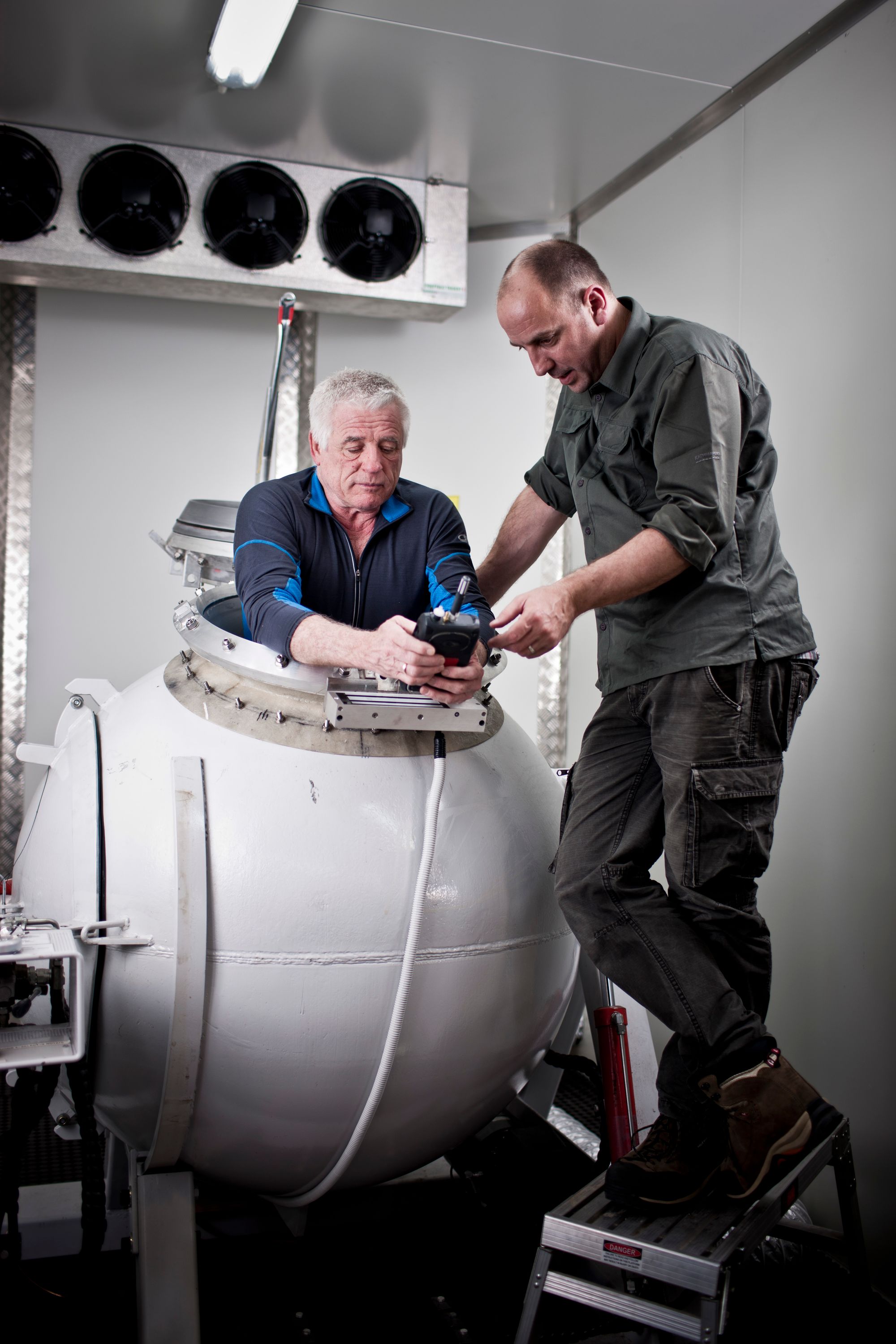 The torso of man with white hair and a blue shirt is visable, while his body is inside a large white sphere. He is talking to another man wearing dark grey and black is standing next to the sphere.