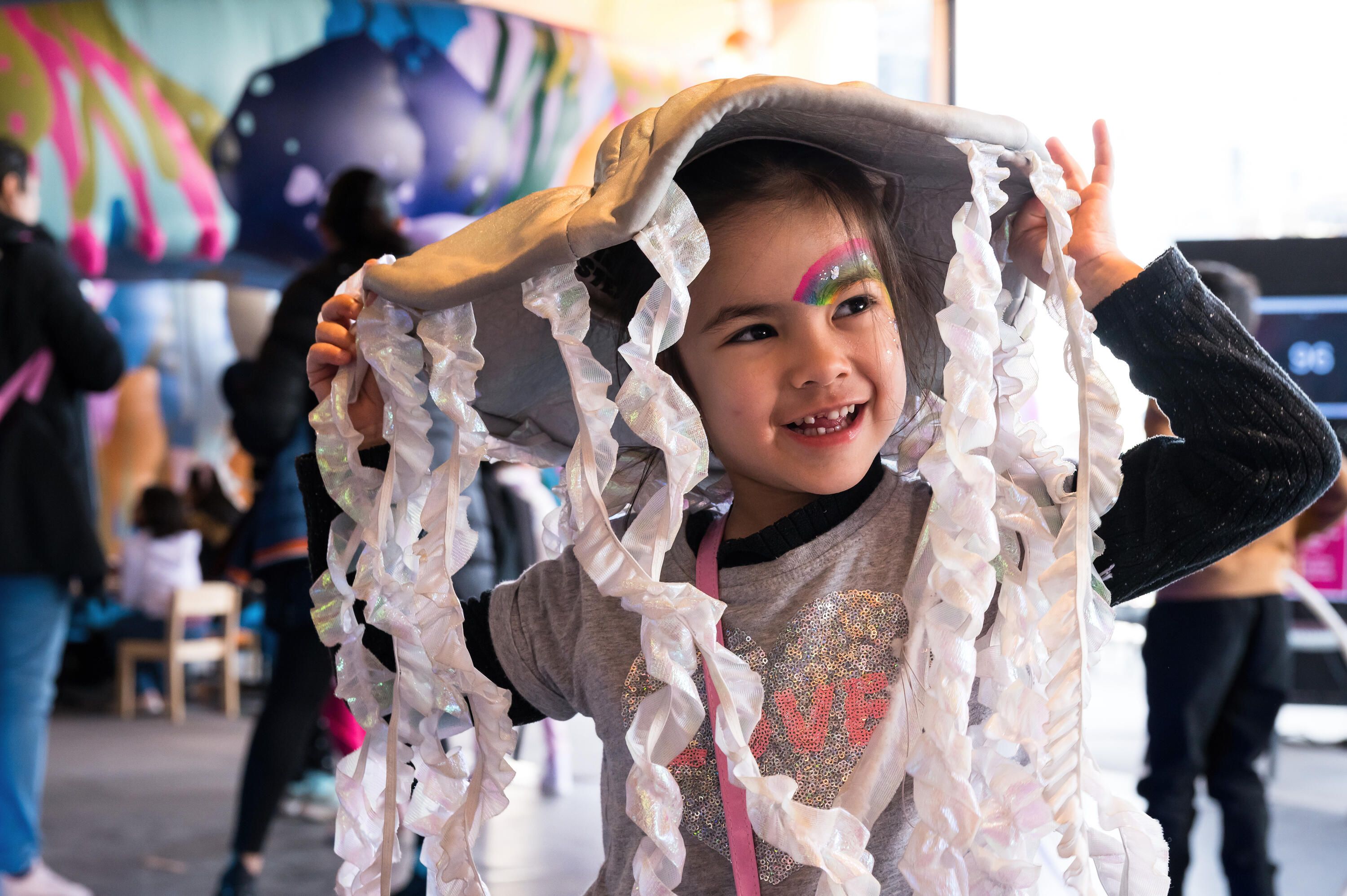 Young girl with a rainbow painted over one eye, wearing a soft jellyfish toy on her head. 