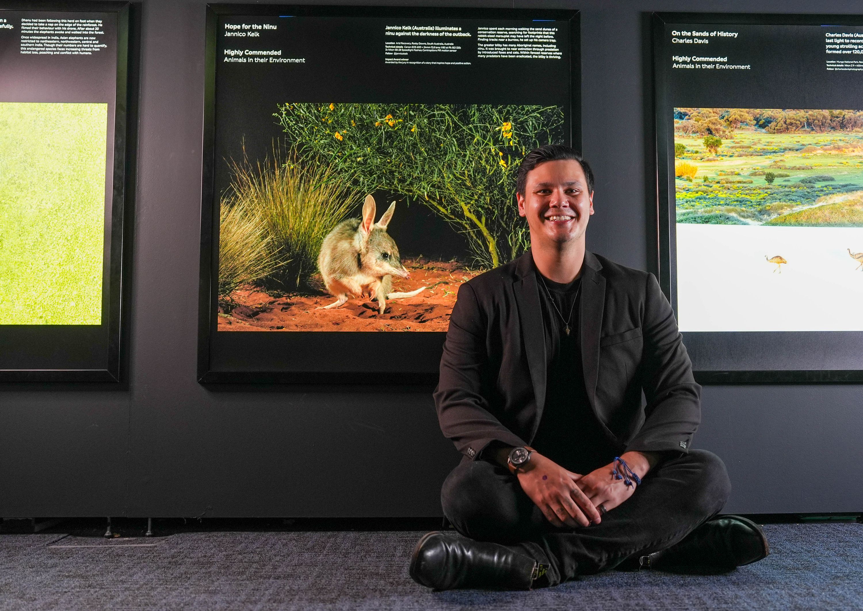 Photograph taken inside an exhibition of a man sitting on the floor next to a photo of a small, fury Bilby.