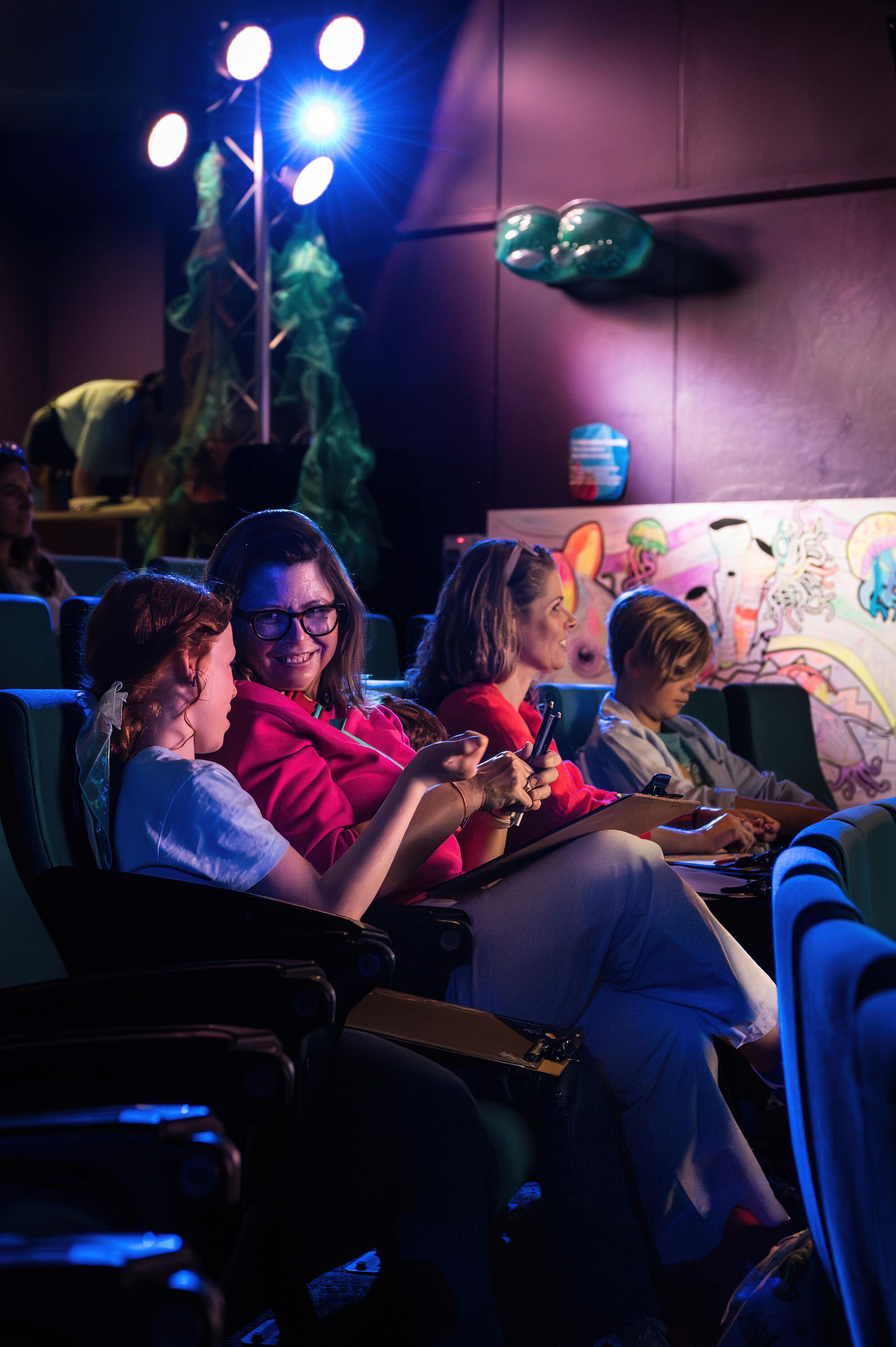 Photo taken inside a theatre showing a girl and an adult smiling, holding a clipboard and pencils. There is another woman and boy behind them.