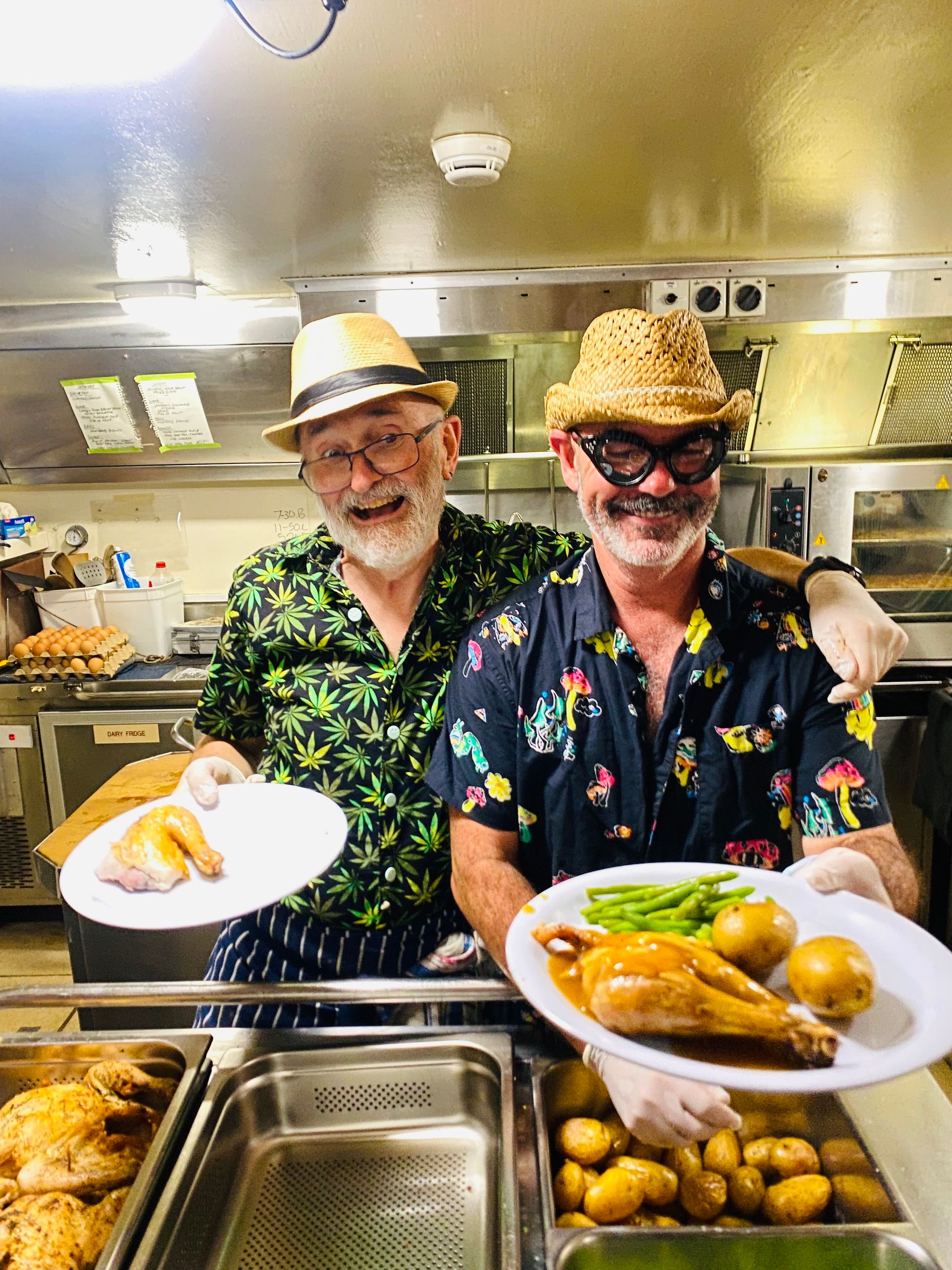2 men in colourful shirt and cream hats holding plates of food in a ship's galley (kitchen)