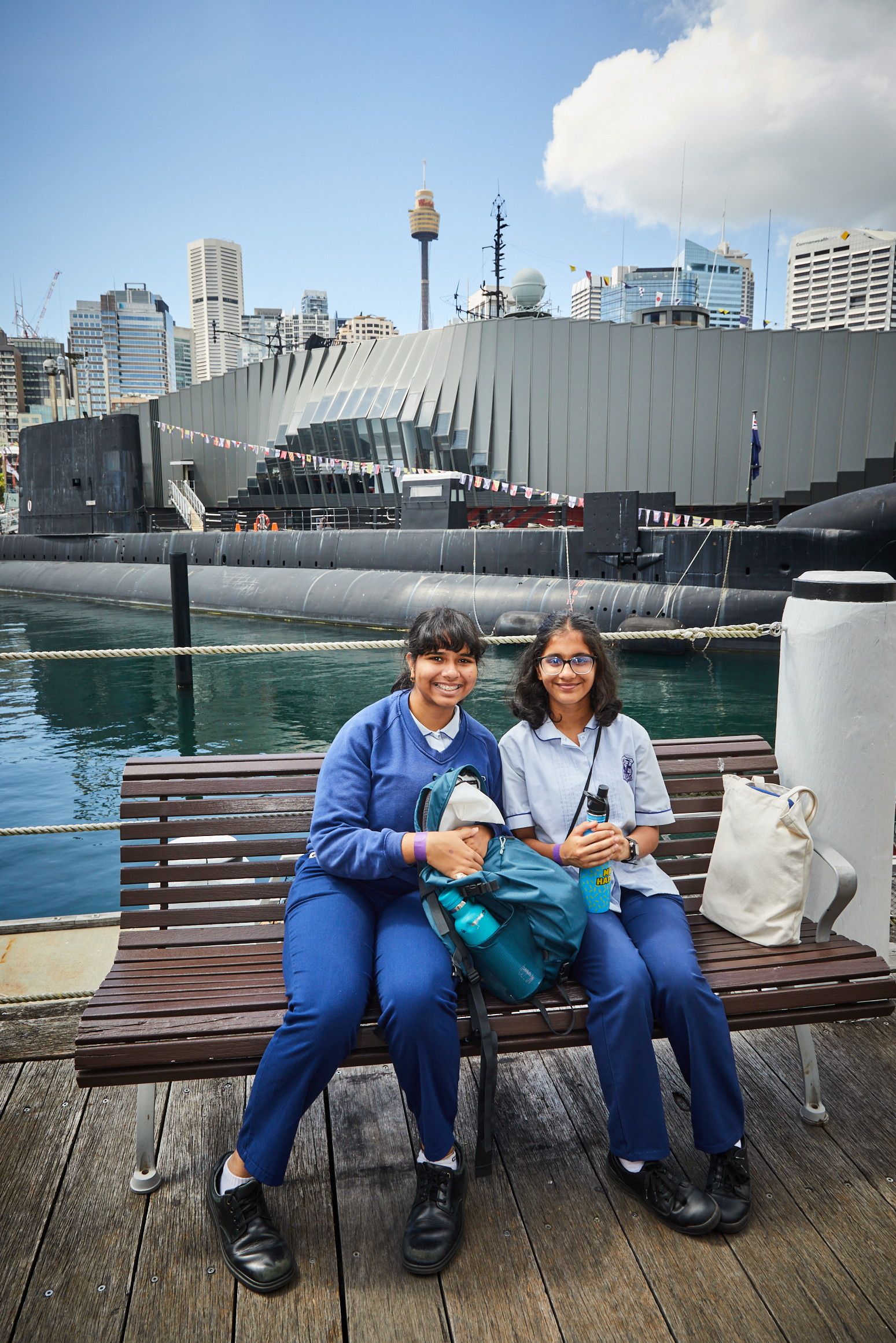 Two girls sitting on a seat outsitde he museum. There is water, a submarine and a building behind them.