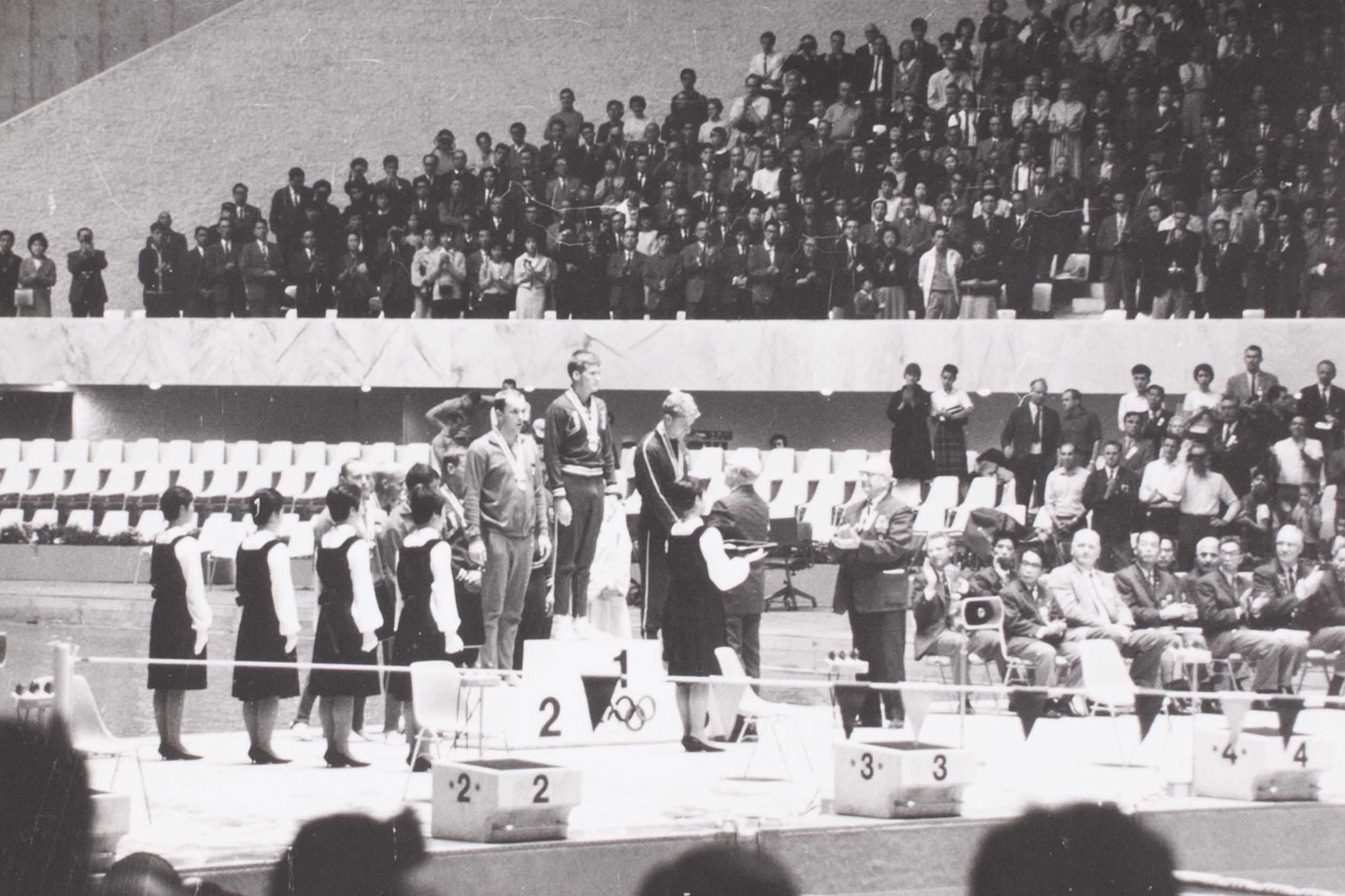 Black and white photo showing a medal podium with people standing on it and a crowd behind them.
