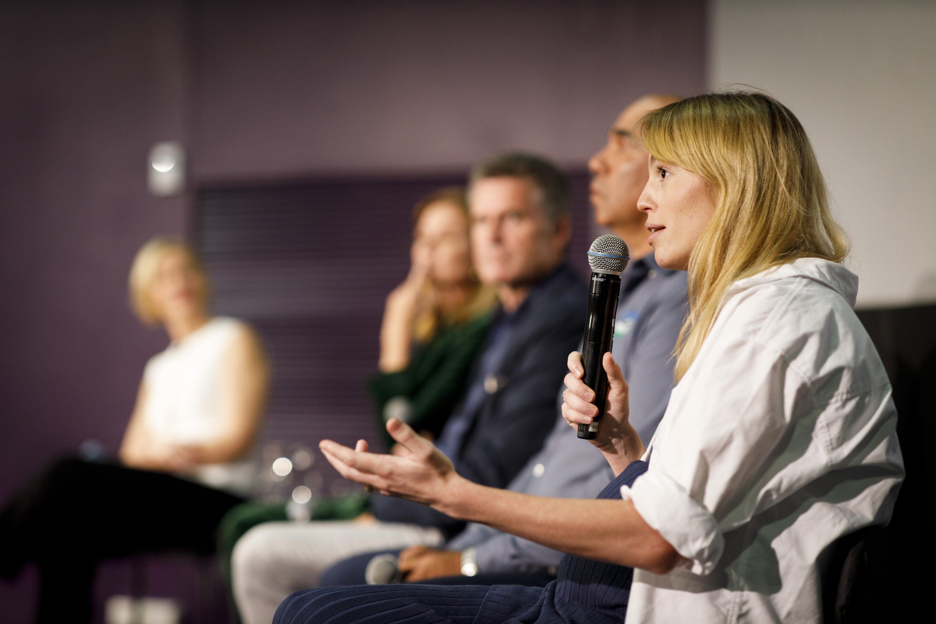 Photo of a woman talking into a microphone, with a line of other people, at the front of a theatre. 