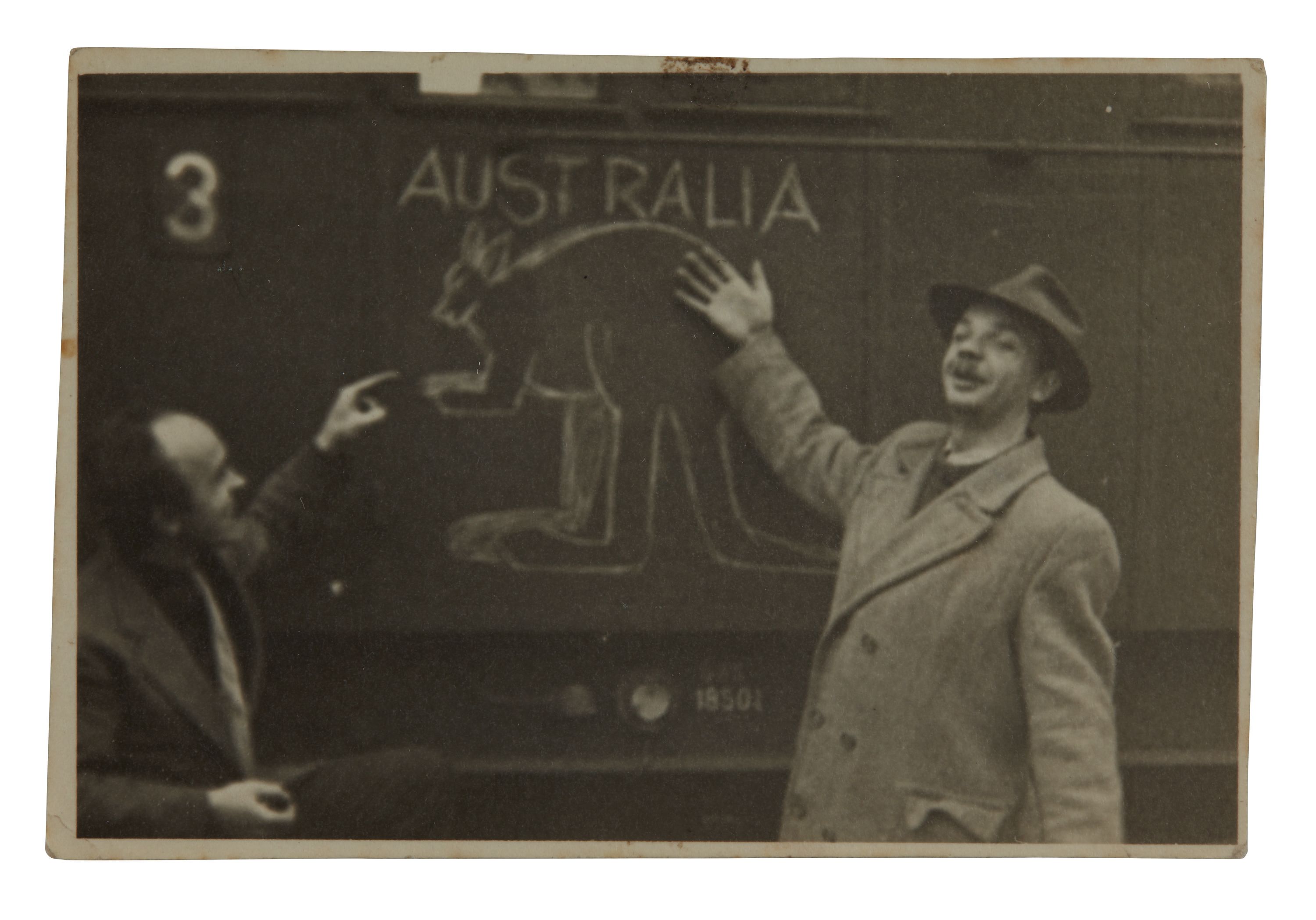 Black and white photo showing 2 men standing next to a wall which has the word Australia and the outline of a kangaroo drawn on it. 