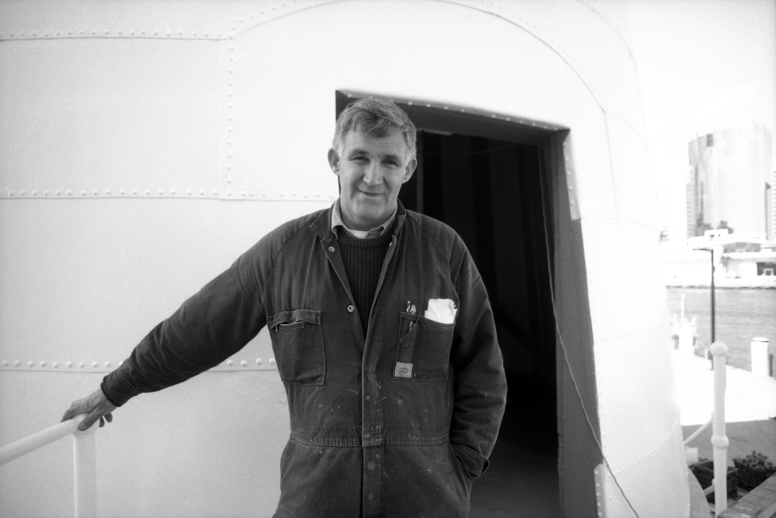 Black and white portrait photograph of a man wearing overalls standing at the entrance to the lighthouse.