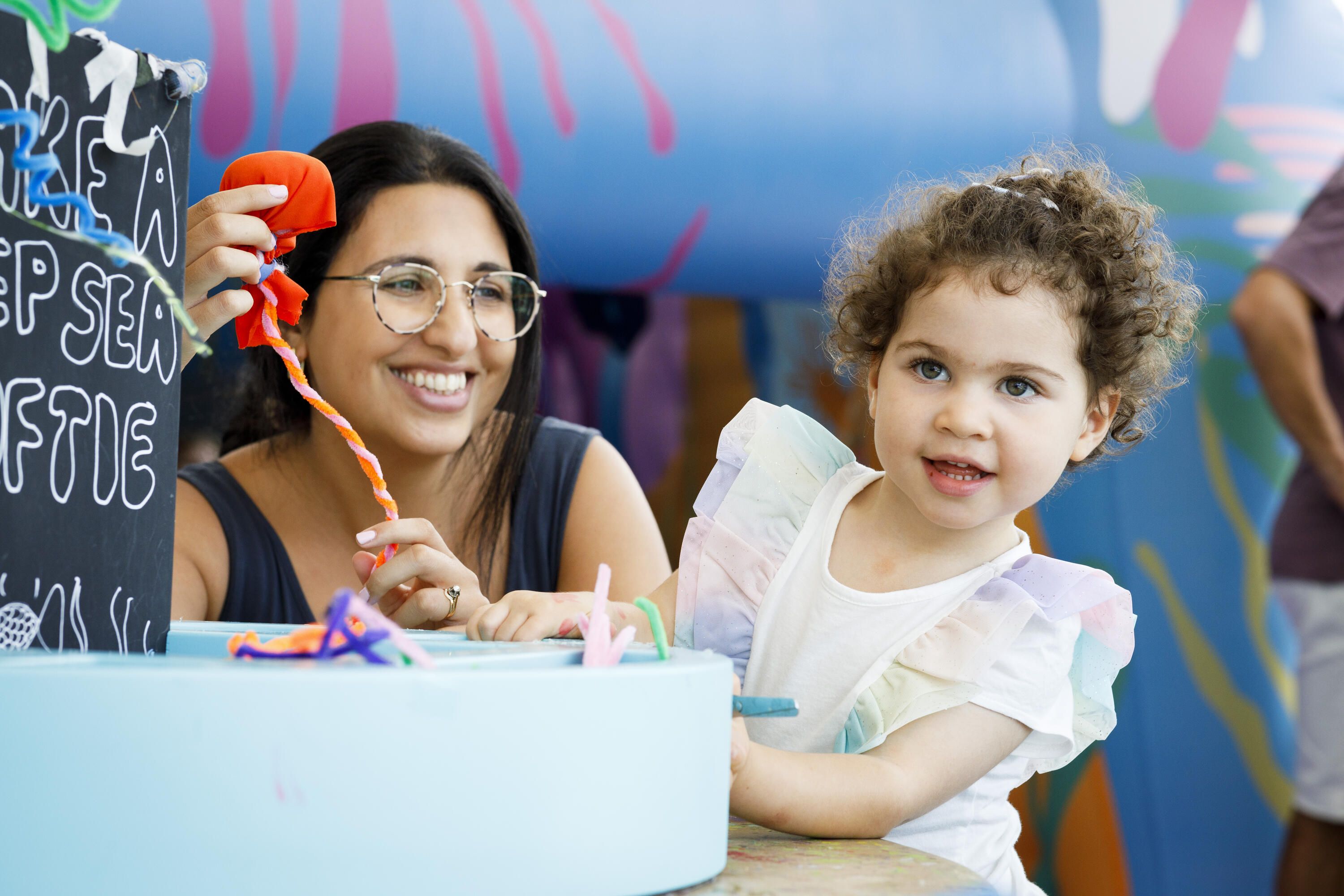 Photo of a child with curly brown hair and a white frilly top next to a woman with black hair, tanned skin, glasses and a black singlet top.
