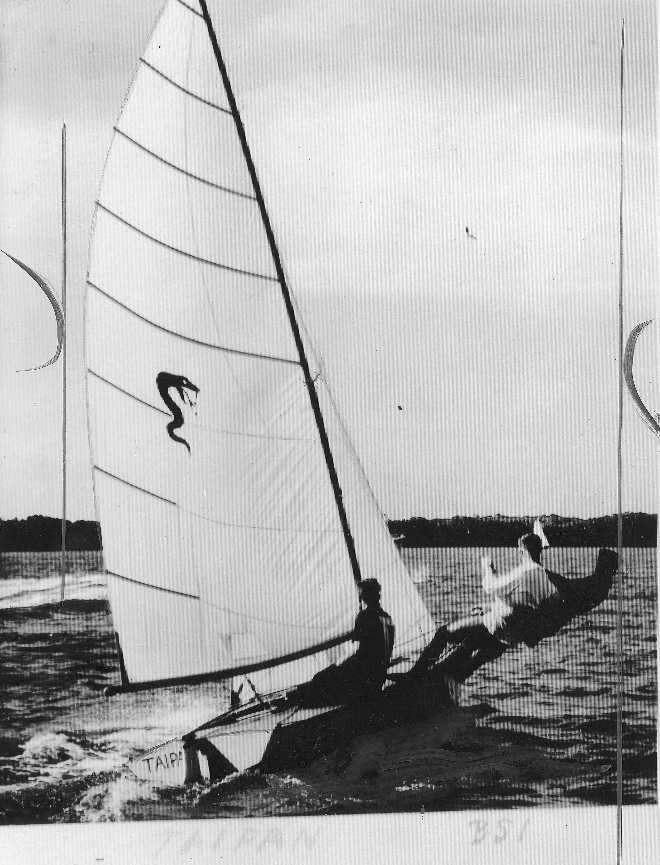 Black and white photograph depicting a three-man crew sailing a sail boat. A black taipan snake motif is visible on the main sail