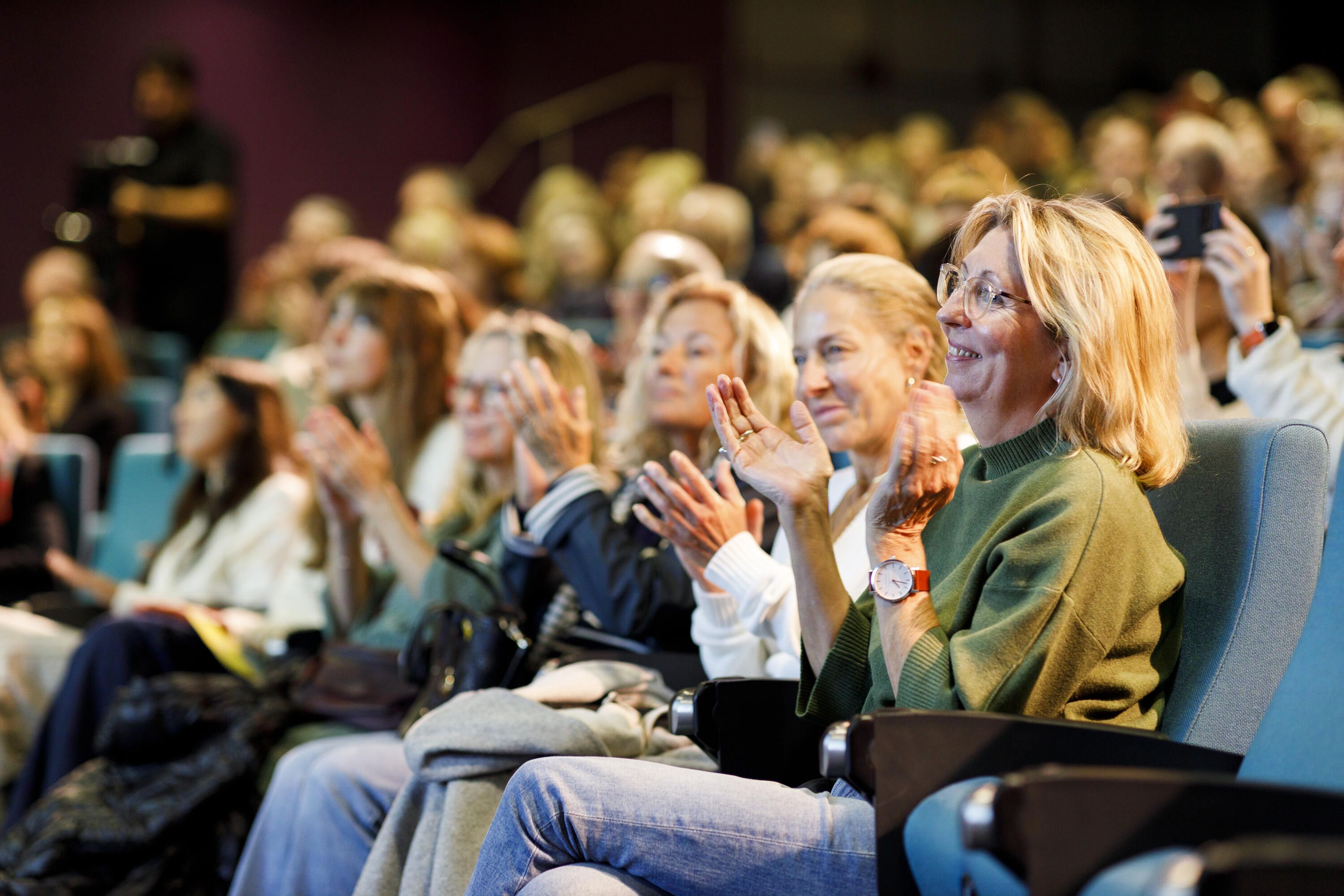Photo of a crowd in a theater. The women in the front row are clapping.