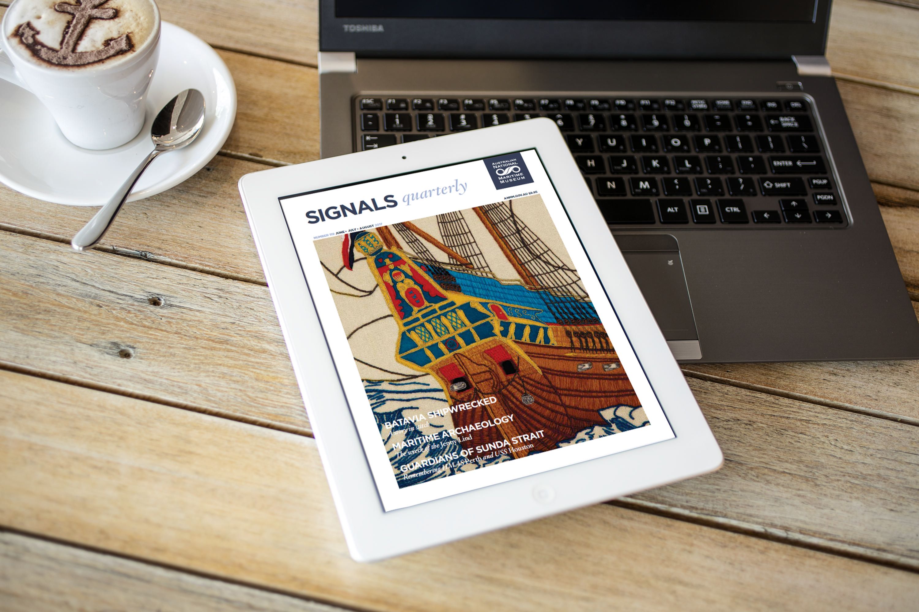 Photograph of a laptop and iPad on a wooden table. The iPad shows the cover of a magazine called Signals. 