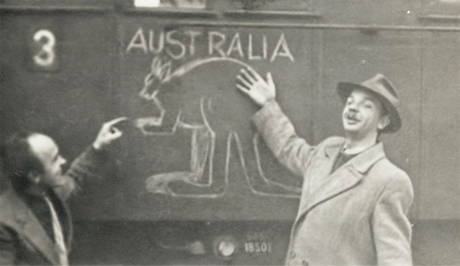 Black and white photo showing 2 men standing next to a wall which has the word Australia and the outline of a kangaroo drawn on it.